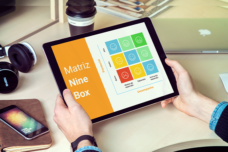 um par de mãos segurando um tablet em cima de uma mesa, com a imagem da matriz nine box na tela, sendo um quadrado laranja na lateral e varios quadrados coloridos em azul, verde, amarelo e vermelho do lado direito